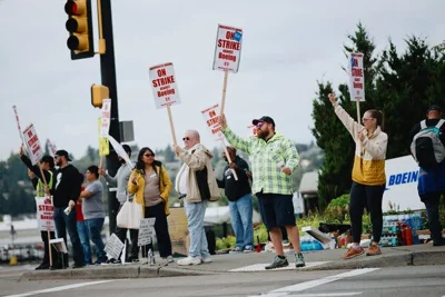 About 10 people standing at a street corner holding signs saying, “On Strike Against Boeing.” A sign with the Boeing logo is behind them.