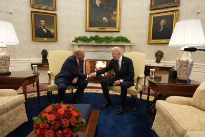 Mr. Biden and Mr. Trump shaking hands while sitting in armchairs in front of a fire. 