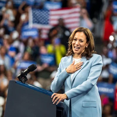 Kamala Harris wears a light blue suit and white shirt. She stands at a lectern with her hand on her chest.