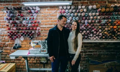 Oleksii Yevsiukov and Viktoriia Varenikova in their clothing factory with a sewing machine behind them and cotton reels on the wall.