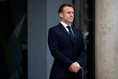 President Emmanuel Macron of France standing in a dark suit and tie with his hands clasped in front of him. 