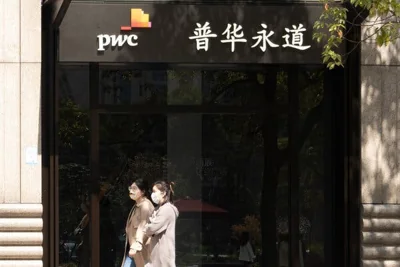 Two women wearing masks walking past an office with a logo for PwC.