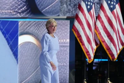 Linda McMahon walks onto a stage decorated with American flags wearing a baby blue dress.