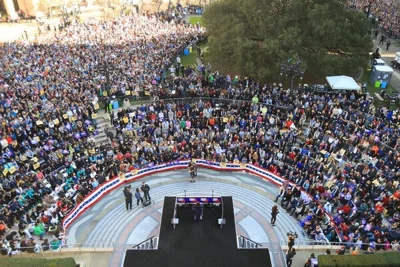 A huge crowd of people ringing the amphitheater outside Oakland City Hall on a sunny day. Kamala Harris stands at a lectern on a platform at the center.