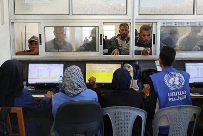 UNRWA workers, one in a blue vest with the UN logo, sit behind glass service windows, behind which is a crowd of people. 