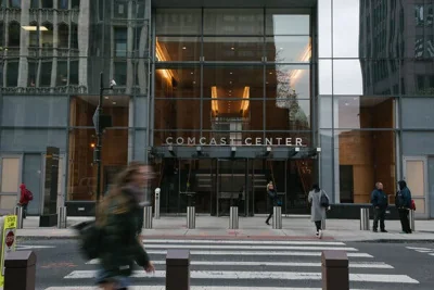 The outside of a steel and glass skyscraper, with a sign saying “Comcast Center” above the revolving door, with a few people walking by near the crosswalk outside.