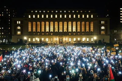 A large crowd of protesters holds up their phone flashlights in front of a Columbia building with illuminated windows.