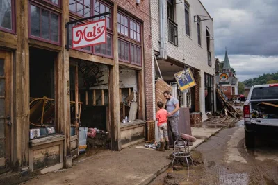 A man and a child stand on a sidewalk along a street covered in mud, with flood damage to nearby buildings. 