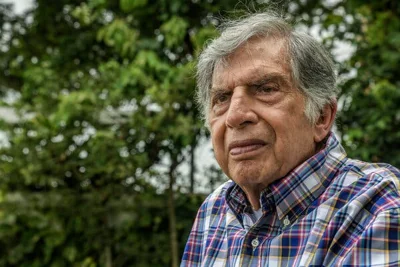 A close-up photo of him against a backdrop of green trees. He is wearing a plaid shirt and has a full head of gray hair and craggy features. 