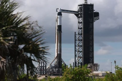 A SpaceX Falcon 9 rocket with the Crew Dragon capsule at the top standing at the launch stand in Florida on a partly cloudy day.