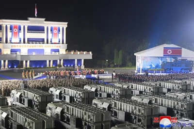 Missile launchers lined up in Pyongyang. They are in rows with troops standing in formation in front. Kim Jong Un is giving a speech from a stage in front of a building. Other officials are standing alongside. The area is floodlit.