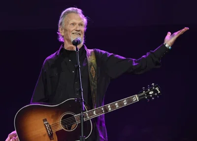 Kris Kristofferson as an older man dressed in black, gesturing on stage in front of a microphone with a guitar hanging over his shoulder.