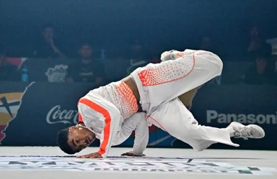 A man in a grey track suit with orange trim balances himself on his hands as he holds his bent legs parallel to the floor.