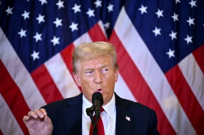 Donald Trump at a news conference. There are US flags behind him. He is holding his right hand up with his finger tips together