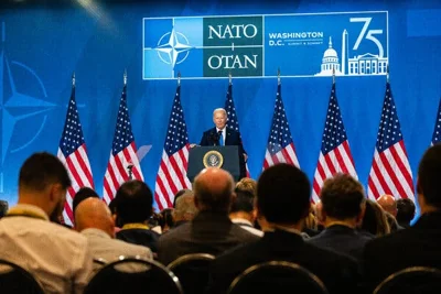 President Biden stands at a lectern in front of a row of American flags and a backdrop with the NATO logo.