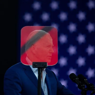 Joe Biden speaking at a rally, his head obscured behind a red-shaded teleprompter. 