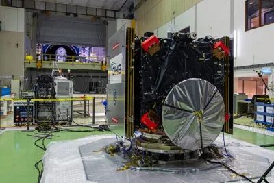 A cube-shaped satellite with its thrusters and antenna dish covered in a testing area of a large space facility.