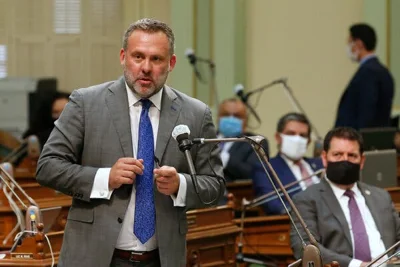 Adam Gray, dressed in a gray suit and blue tie, stands at a microphone.