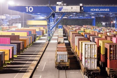 Lines of shipping containers, some on flatbed rail cars, with a series of cranes overhead.