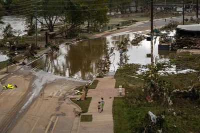 A flooded road seen from above, with a river in the upper left corner and two people standing on a sidewalk. 