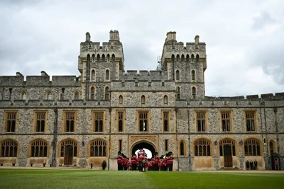 A marching band of soldiers in traditional red uniforms and black fur hats, walking through a gate at Windsor Castle.