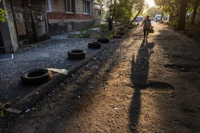 A figure silhouetted against the morning sun casts a long shadow over a road, its sidewalk covered in glass fragments.