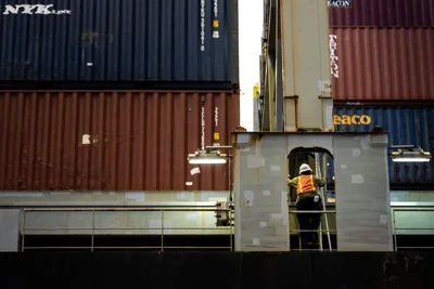 A worker in an orange vest and hard hat on a piece of equipment next to large stacks of maroon and blue containers.