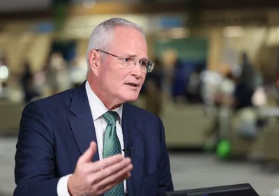 Darren Woods, wearing a navy blue suit, a white shirt and a green tie, is sitting and holding up his right hand.