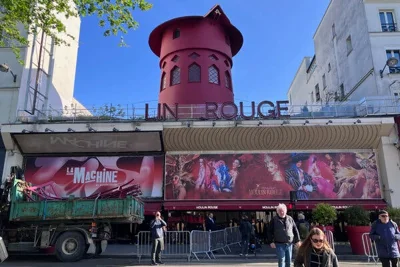 The front of the Moulin Rouge, with a green truck in front of the entrance carrying bent pieces of a red windmill that fell from the building. 