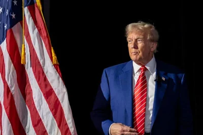 Former President Donald J. Trump, wearing a blue suit and red striped tie, stands next to an American flag.