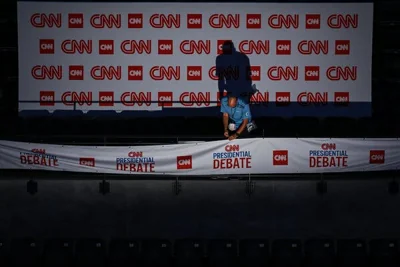 A man in a uniform fixes a sign that says CNN Presidential debate, with his shadow on the wall behind him, which has many more CNN logos.