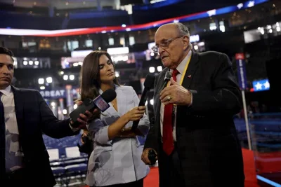 Rudolph W. Giuliani, wearing a dark suit and red tie, speaking with two people holding microphones close to him.