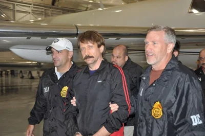 Viktor Bout accompanied by US law enforcement officers. He is dressed in black and has a large moustache.