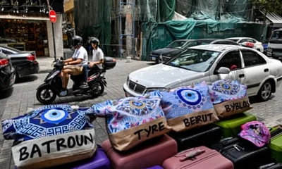 In the foreground, beach bags displaying the names of the Lebanese cities of Batroun, Tyre, Beirut and Byblos sit on suitcases, with two men on a motorbike and a car in the background and, to the right, a bombed building covered in green netting