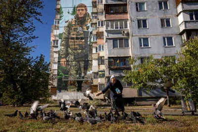 A woman feeding pigeons in front of a mural on the side of a building of a Ukrainian soldier.