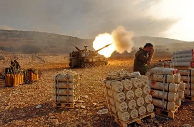 A large flash erupts from an armored vehicle on a barren hillside as it fires a shell.  A soldier in the foreground, surrounded by unused shells, covers his ears.