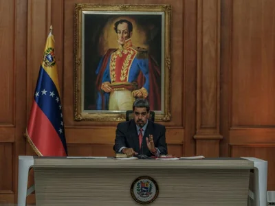 President Nicolás Maduro sitting at a long desk in a suit with a flag to his right and a portrait of the military leader Simón Bolívar behind him.