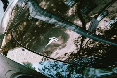 A building is reflected onto the hood of a black Tesla vehicle, with the Tesla logo showing.