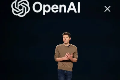 Sam Altman speaks on a stage. A screen with the OpenAI logo appears behind him.