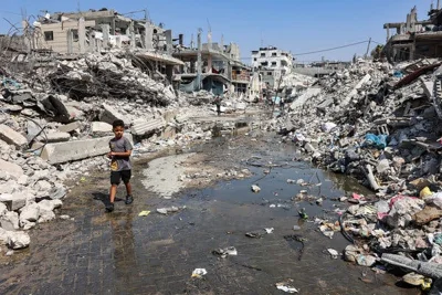 A boy wearing a gray T-shirt walking through a puddle of sewage water and past mounds of trash and rubble.