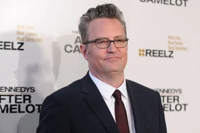 Matthew Perry wears glasses while standing in front of a white backdrop.