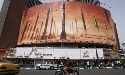 A huge billboard in central Tehran shows Iranian missiles: they are depicted as vertical ballistics against an orange background on a curved billboard in a city street, as cars, trucks and motorcycles pass
