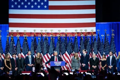 Donald J. Trump standing onstage under an enormous American flag, his arms spread, with a row of people and American flags behind him.