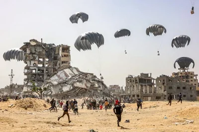 A wide view from the northern Gaza Strip of people running toward humanitarian aid packages arriving by parachute. In the background are bombed-out buildings.