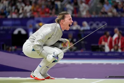 Olha Kharlan, in a white fencing suit, crouches as she celebrates, holding a saber.