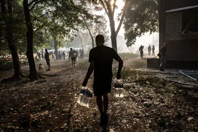 A silhouette of a man holding water bottles in Ukraine. A crowd is gathered near trees in the background.