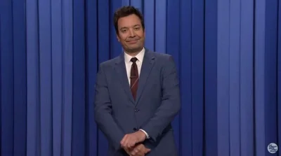 Jimmy Fallon, in a gray suit and burgundy tie, stands onstage in front of a blue curtain.