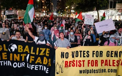 A crowd of people, some wearing kaffiyehs and holding Palestinian flags or banners, fills a street.