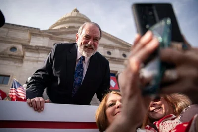 The former Arkansas governor Mike Huckabee, in a suit, leaning forward as a woman takes a selfie with him. The Arkansas State Capitol is in the background. 