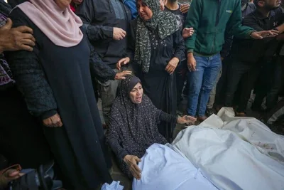 A woman sits on the ground next to three bodies wrapped in white. People stand around her.